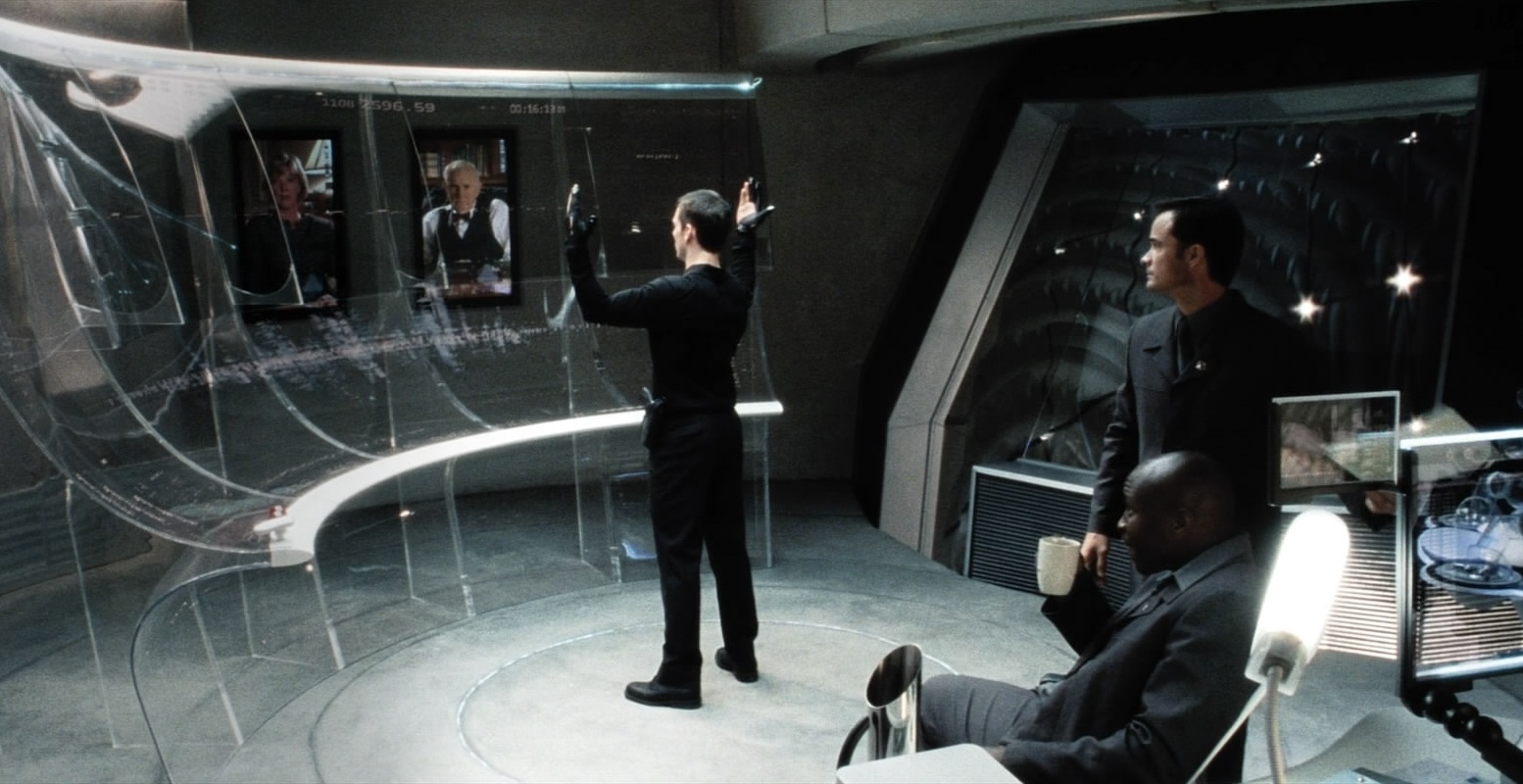 Tom Cruise, vestido inteiramente de preto, gesticula em frente a uma tela curva e transparente, observado por outros dois homens.