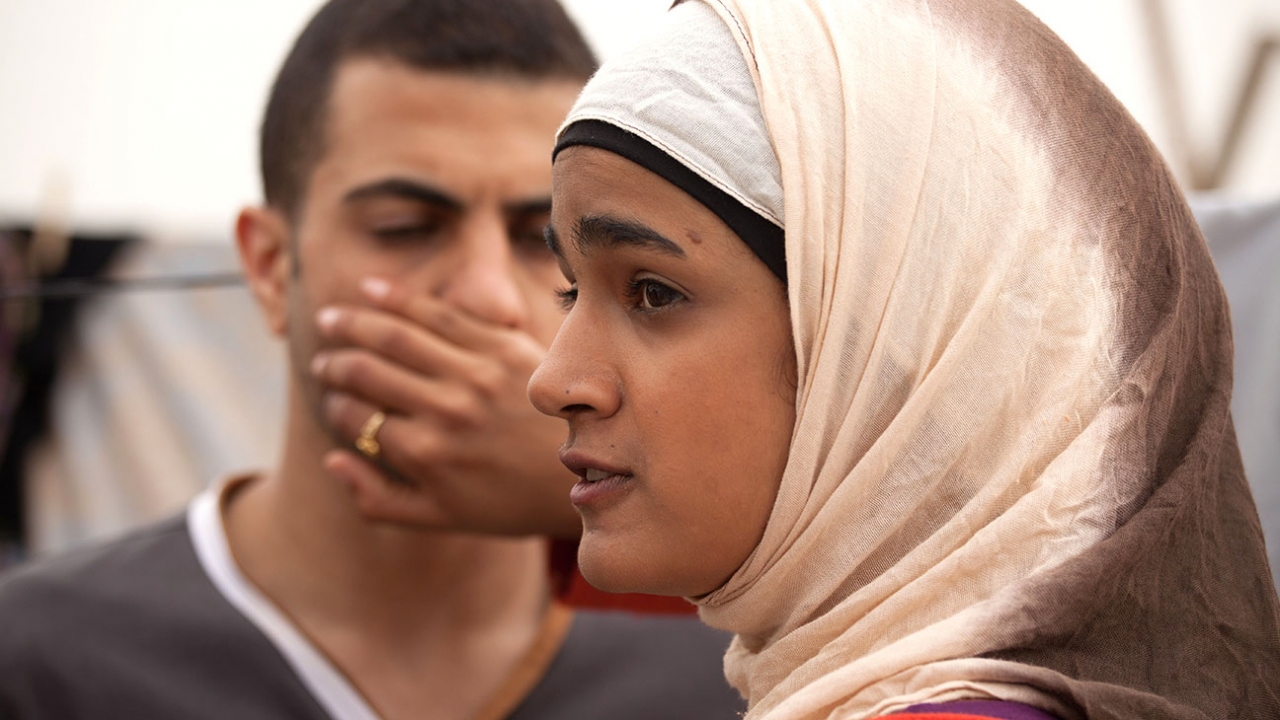 Uma jovem de hijab rosa clarinho e camiseta de manga longa rosa pink, cobre a boca de um rapaz que a observa com sua mão. 