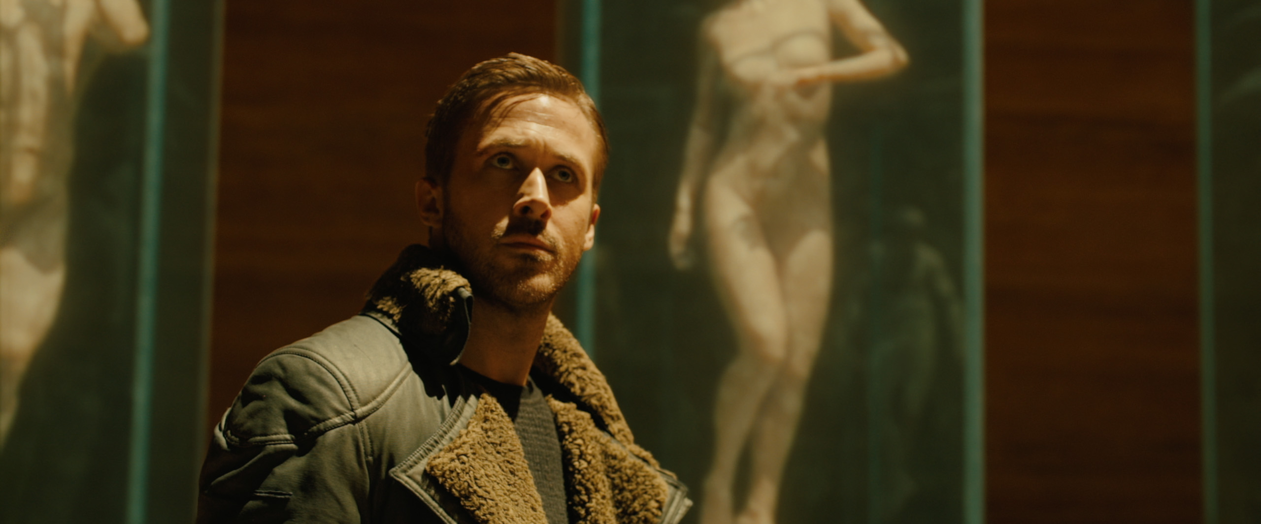 Ryan Gosling, vestindo uma jaqueta verde com lapela larga de pele falsa de carneiro. Atrás dele uma espécie de aquário mostra um corpo feminino nu sendo gerado.