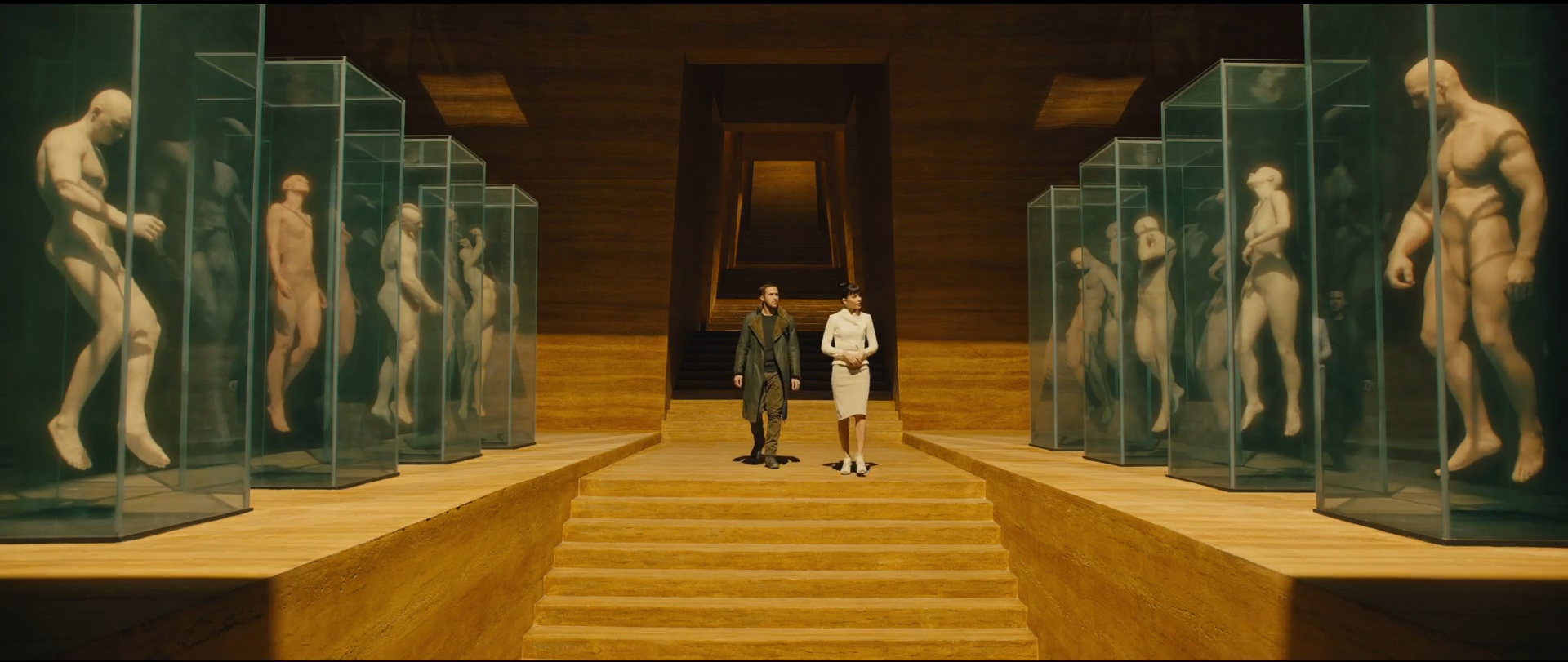 Rya Gosling e Sylvia Hoeks (Luv) caminha em direção a uma escada em um grande salão rodeado por aquários contendo corpos humanos