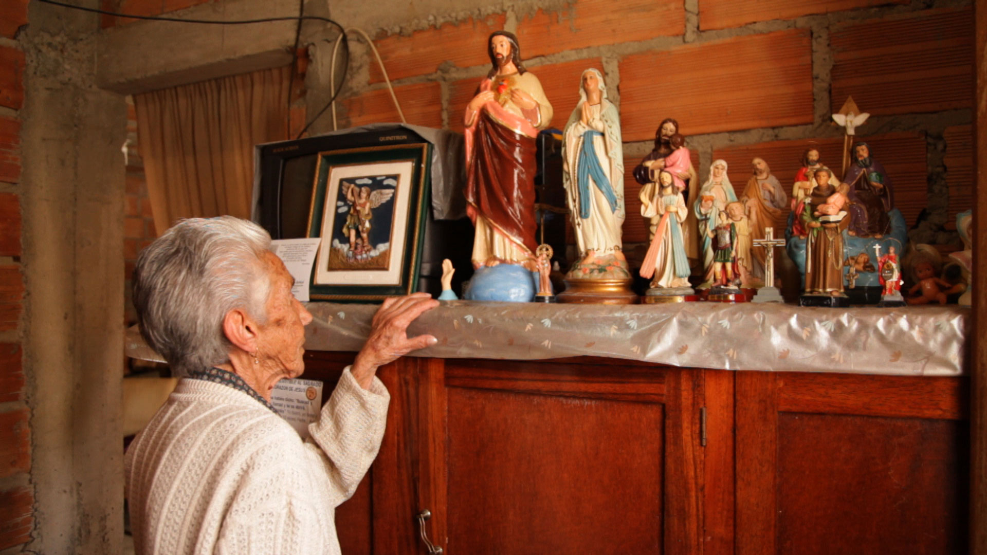 Uma senhora de costas, com cabelos brancos, olha para uma coleção de esculturas de santos perfiladas sobre um armário