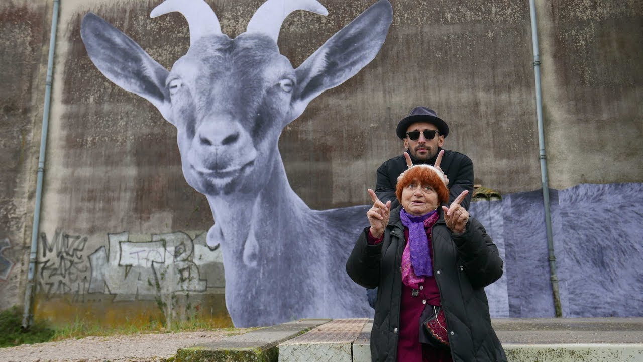 Um grande muro com uma foto gigante de um cabra colada sobre ele. Na sua frente, Agnès Varda e JR, diretores do filmes, fazem chifrinhos com os dedos
