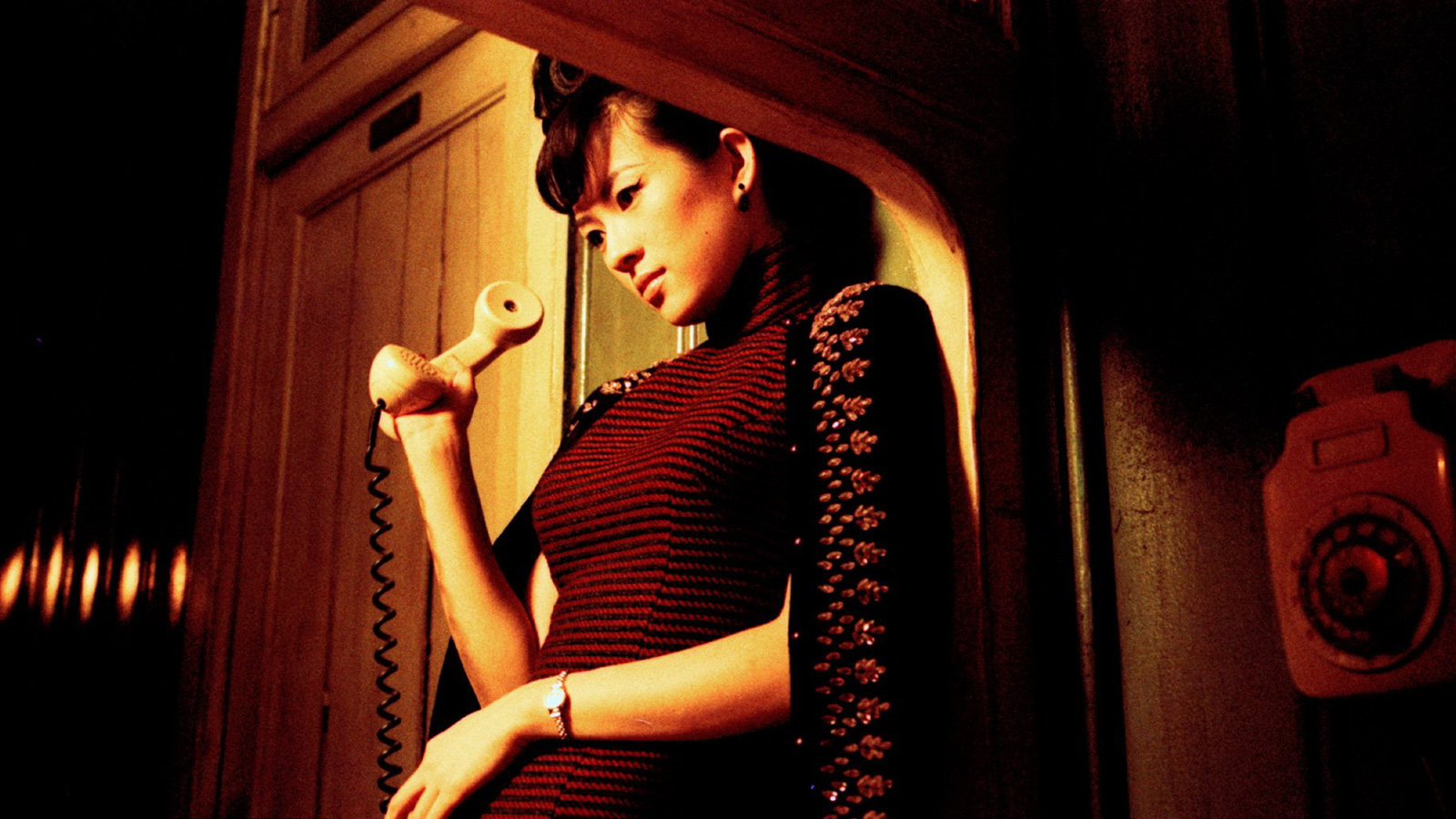 A atriz Zhang Ziyi, de 2016, apoiada com as costas em uma parede segurando um telefone na mão enquanto olha o chão. usa um vestido justo e listrado em tons escuros, um casaco preto sobre os ombros e um relógio de pulso pequeno com pulseira fina no pulso esquerdo. 